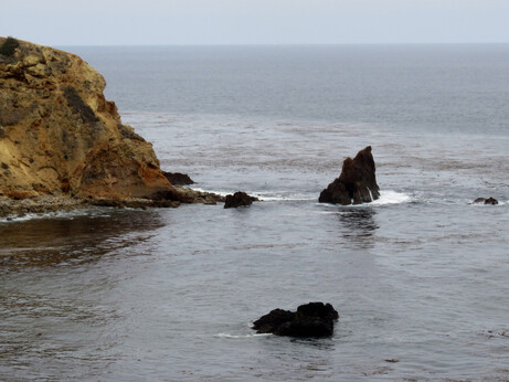 Jagged rocks out in the smooth ocean, not far from a rocky cliff, on a gloomy day.