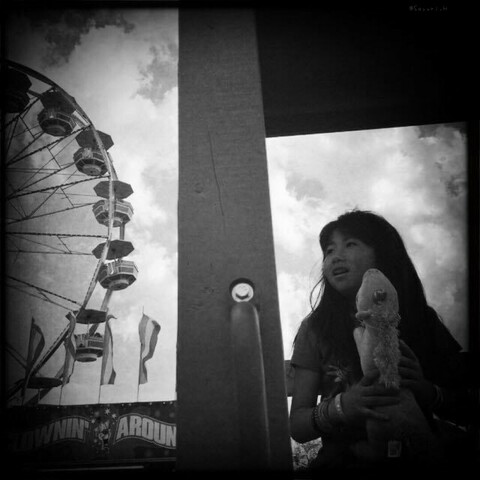 Black and white photo of a girl holding unicorn stuffed animal that she won as a prize.  She’s sitting next to ferris wheel