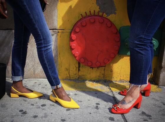 2 woman legs, they one is wearing yellow shoes and the other red shoes. The background wall is red and yellow