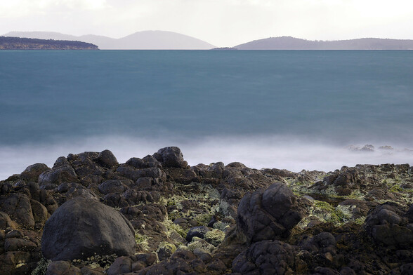A long exposure of a rocky foreshore looking out across a large bay, with seaweed amongst the rocks in foreground.  The foreshore boulders catch the sunlight coming from the right and the bright patchy blue sky (not visible) reflected on the left, creating an almost surreal lighting.  The sea is a dark turquiose from the long exposure and diffused light created by almost full cloud cover.  The land features and hills in the distance are progressively more grey as the rain approaches.