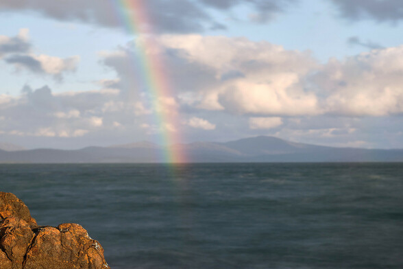 A view across a large bay towards distant hills, with a rainbow from close in the foreground rising out of view. The water surface is smoothed by a long exposure, a dark greenish blue.  An orange brown rocky outcrop appears in sharp focus at the lower left.  The sky is a patchy pale blue with low level cloud covering most of the sky.