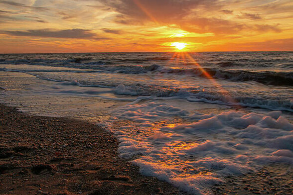 Sunrays appear at the horizon over the Atlantic Ocean while foaming waves wash up on the beach in Cape May, New Jersey. The sunset is a glowing orange.  