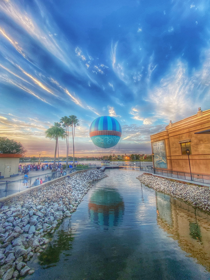 A balloon landed at the edge of the lake and you can see the reflection of the balloon on the water. The sky is changing colors to orange as it’s getting late in the evening. People are gathering by the dense to look at the balloon