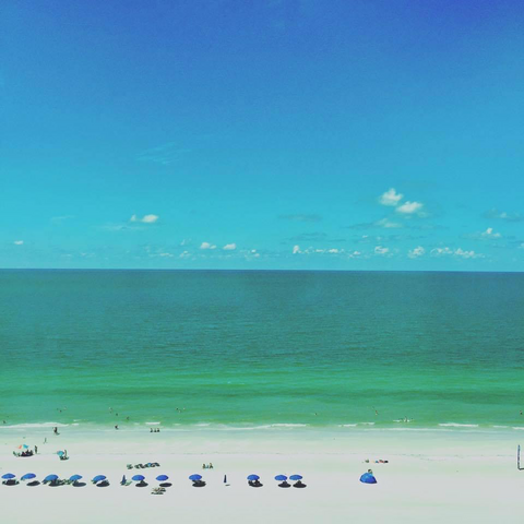 Emerald green water, white sand, and blue sky at Naples beach. Blue beach umbrellas lined up on the sand 