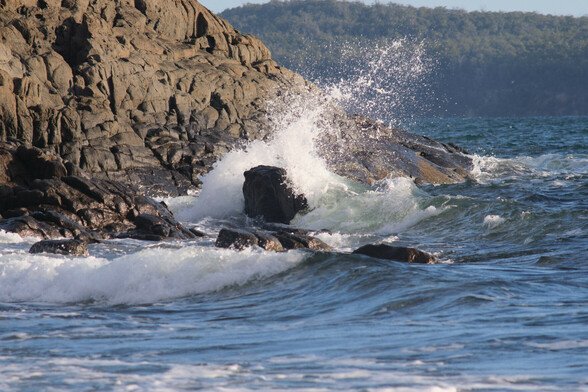 A wave crashing over a rock next to the shoreline, the force reflecting the power and sending the spray backwards.  The shoreline looks like one large piece of weathered rock, which curves around a point.  Tree lined hills fill the horizon across the bay at right.