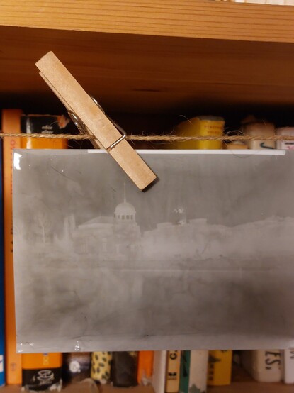 Still wet photo, hanging on a thread and fastened with a clothespin. It is a flipped negative of a view of an old hydroelectric power station, situaded on an island of a river. The photo is blurry and has many defects, such as swirls of uneven color. 