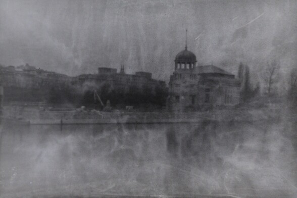 Processed photo made from a flipped negative. It is a view of an old hydroelectric power station, situaded on an island of a river. The photo is blurry and has many defects, such as swirls of uneven color. 