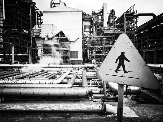 A black and white photo of an oil refinery. The photo shows a series of pipes, tanks, buildings, and in the foreground is a warning sign with a man on a foot path showing the location of the foot path.