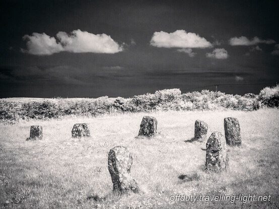 View of a part of a stone circle. Beyond the circle is a hedge and clear black sky with a line of small white clouds.