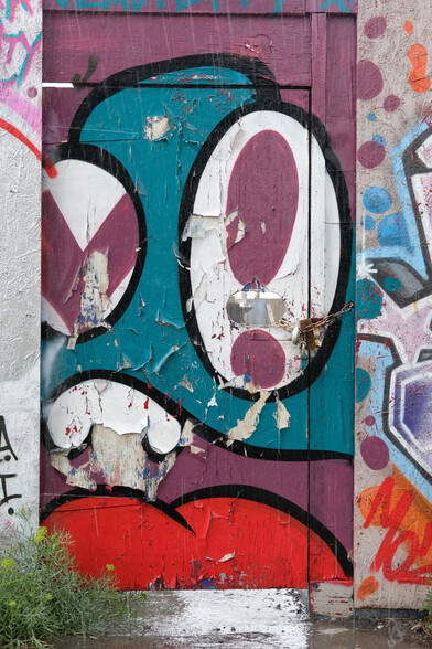 A colour photograph, taken in a down pour of rain, of a OSB woodflake door which has been grafittied over - several times now.
[The street artwork is an abstract face of sorts.] 