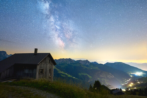 Mountain landscape with Milky Way in the sky. On the left is a mountain hut in the foreground and on the right are the lights of the valley.