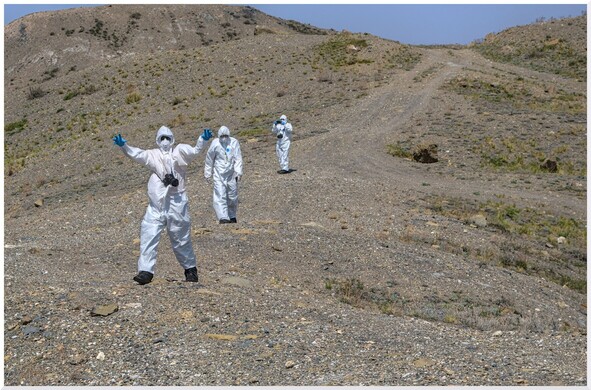 A group of three people.in hazmat suits walk across a gravel road on the Kazakh steppe. The one Infront has arms out stretched like a zombie