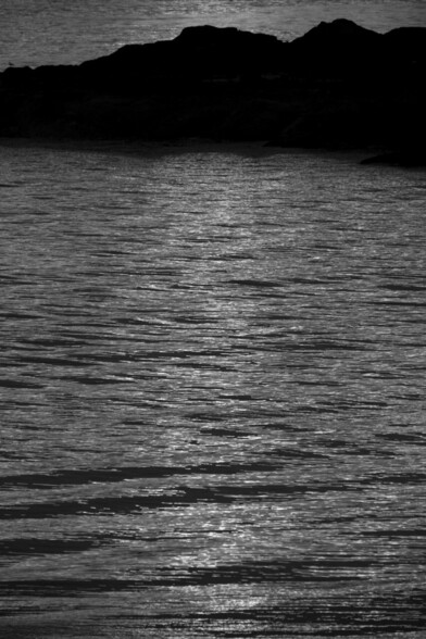 Black and white portrait format image taken at sunset and showing the light from the setting sun creating a channel across the gently rippling ocean waves, while in the background is the stark black outline of coastal rock formations