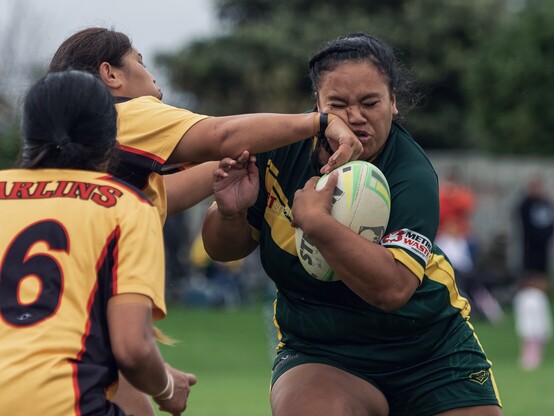 A rugby league player’s face contorts as she gets an opposing player’s wrist in her face as she runs into a tackle.