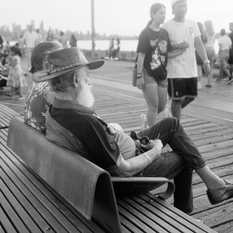 A gentleman sits on a bench on the pier and watches the crowd roll by.