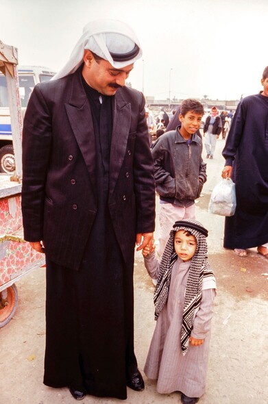 Vertical portrait of father and very young son, probably 3-4 yo, hand in hand and in traditional Arab clothes. Father is smiling and looking down at son. White sky, diffuse light with almost no clouds.