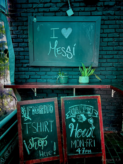 The green bricks wall has blackboard sign that says “I love Messi”. There’s a thin and long wood shelf with two pots of plants. There’re two blackboards that are standing on the floor 