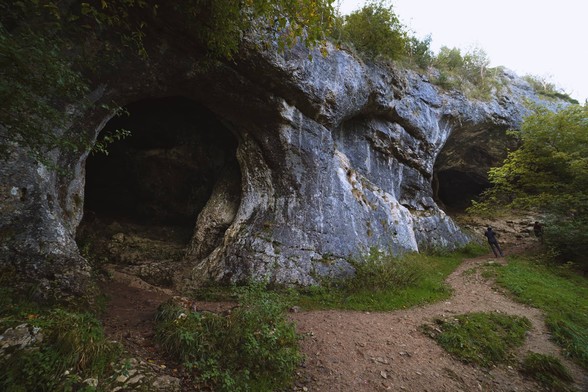 Looking up at two deep and dry caves, covered in foliage around them, with two walkers looking up at them.