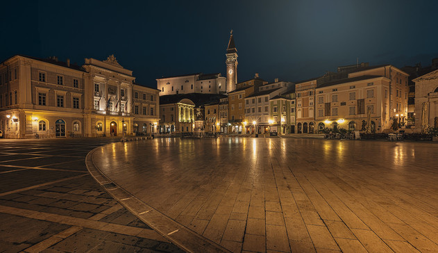 A nighttime view of a classic European square in Piran Slovenia. There are buildings ringing the wide open square. 