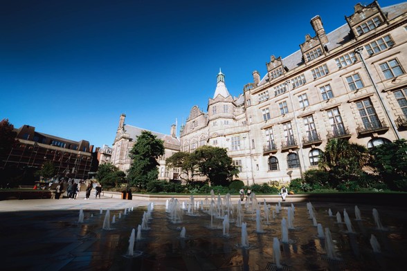 A clear blue sky day in autumn. Below, in the shadow of surrounding buildings, water fountains bubble up from the ground, pointing towards the grand gothic town hall.