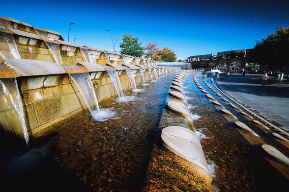 A clear blue sky day. Ultra-wide photo of a series of constructed cascades that curve away towards a railway station in the distance.