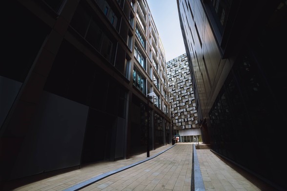 A narrow walkway in between two dark and tall skyscrapers, lining the way towards an illuminated building with a cheesegrater like exterior.