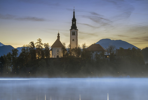 Early morning view of Lake Bled island in Slovenia with the spires of the church and other buildings silhouettes against the dawn sky. Fog rising from the lake in the foreground. 