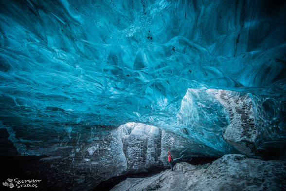 Inside an ice cave under a glacier with blue translucent ice above. A person in a red jacket standing beneath two openings in the ceiling where light comes in