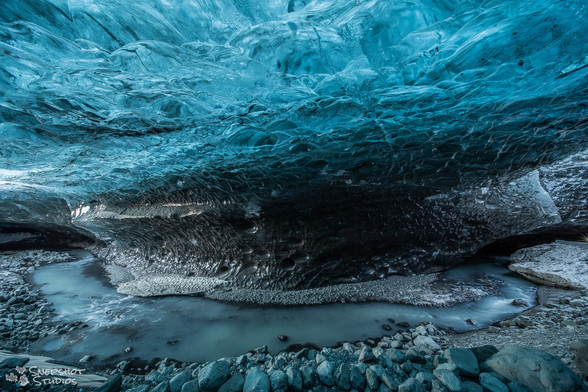 Inside an ice cave under a glacier with blue translucent ice above. A small river of melt water curving from right to left.