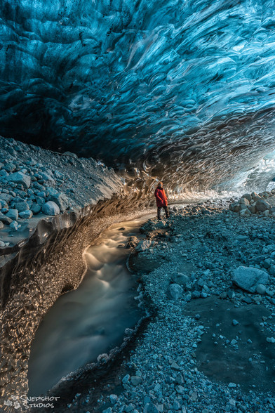A cave in the ice below a glacier. Cold blue light comes through the ice-ceiling. Warm light from an entrance illuminates a small stream of water along the floor and a person in a red jacket. 