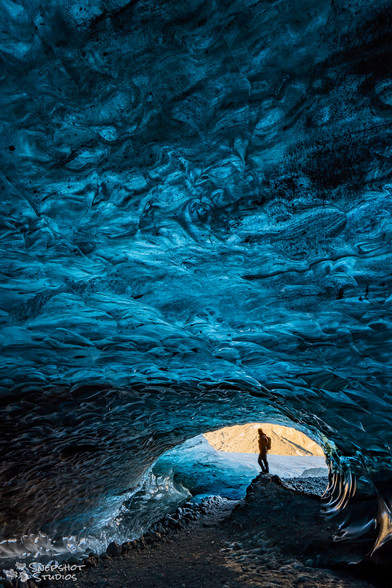A cave in the ice below a glacier. Cold blue light comes through the ice-ceiling. Warm light from an opening in the background illuminates a person in a red jacket. 