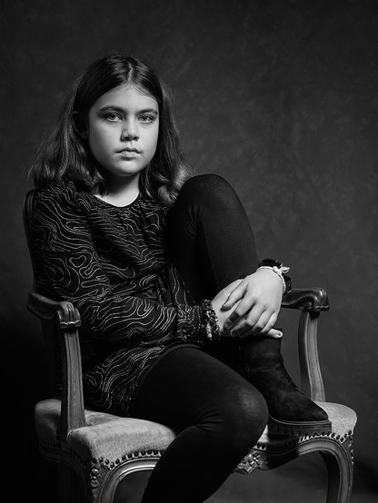 10 jähriges Mädchen, das ernst in die Kamera schaut. Sie sitzt auf einem Sessel, hat ein Bein auf die Sitzfläche aufgestellt und umklammert es mit ihren Händen. Sie hat ein dunkles Kleid, Leggings und Stiefel an. Ihre Haare sind schulterlang, leicht gewellt und schwarz.