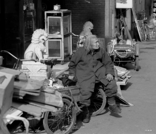 An elderly woman in traditonal chinese clothes is sitting on a cart near some stone sculptures - they seem to resemble each other.