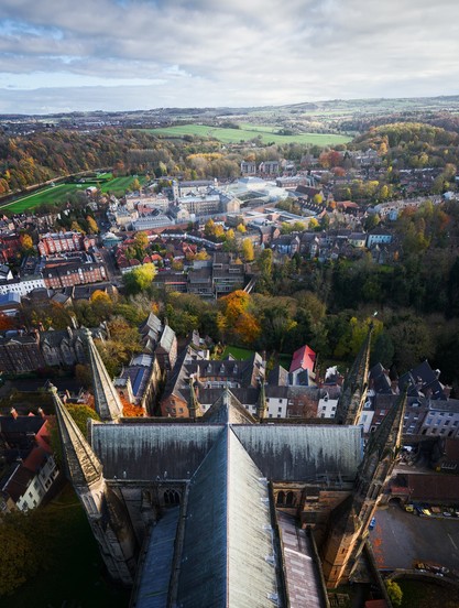 The image is an aerial view of a city with a large cathedral prominently featured. The cathedral has a pointed roof and is surrounded by a town with many buildings. The city is situated in a valley, with a lush green field nearby. The scene is bustling with activity, as there are several cars and a truck visible throughout the image. The overall atmosphere is that of a vibrant and thriving urban environment.
