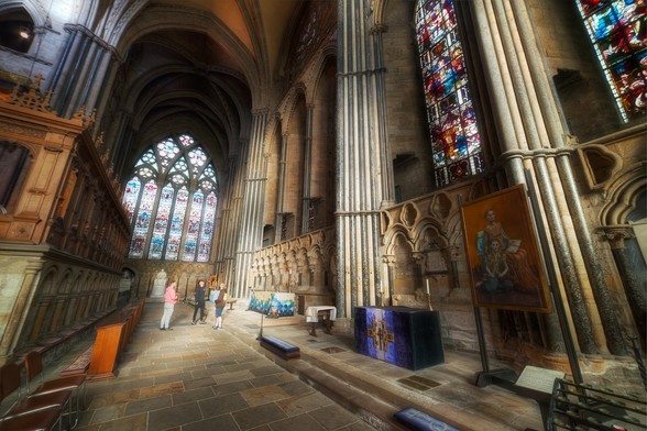 The image depicts a large, ornate church with a spacious interior. The church features a high ceiling and a large stained glass window, which allows natural light to fill the space. There are several benches placed throughout the church, providing seating for the congregation. In the scene, a group of people can be seen walking around the church, possibly attending a service or exploring the space. Some of the individuals are closer to the foreground, while others are further back in the scene. The overall atmosphere of the church is serene and inviting.