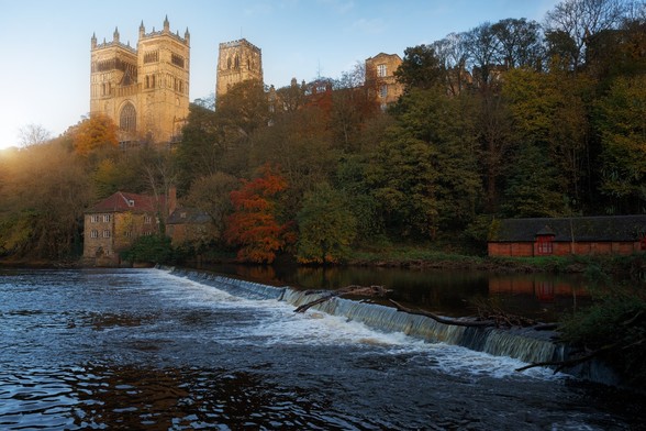 The image features a picturesque scene of a river flowing through a town, with a castle-like building in the background. The river is surrounded by trees, creating a serene atmosphere. The water is rushing, and there is a waterfall nearby. The scene is beautifully captured during sunset, casting a warm glow over the landscape.