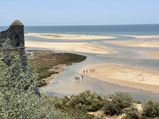 Paysage côtier avec une rivière se jetant dans la mer, un banc de sable, des gens qui marchent, et une section d'un ancien mur de forteresse avec de la verdure.