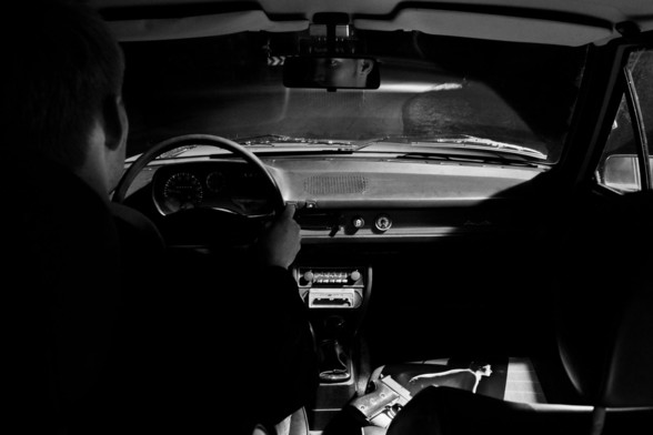 Der Mann sitzt wieder im Auto und fährt zurück. Die Waffe liegt auf einem Bild auf dem Beifahrersitz