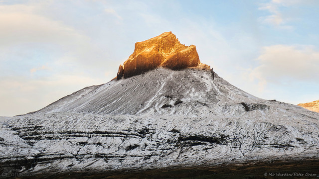 In the centre of the photo is a triangular mountain topped by a strange, steep-sided spike of rock. The flanks of the mountain are covered in snow, all the way up to this unusual peak. The sky behind is blue with some sunset-tinted cloud, and the whole mountain is in shadow with the exception of the very tip, which is highlighted by the colour of the sunset.