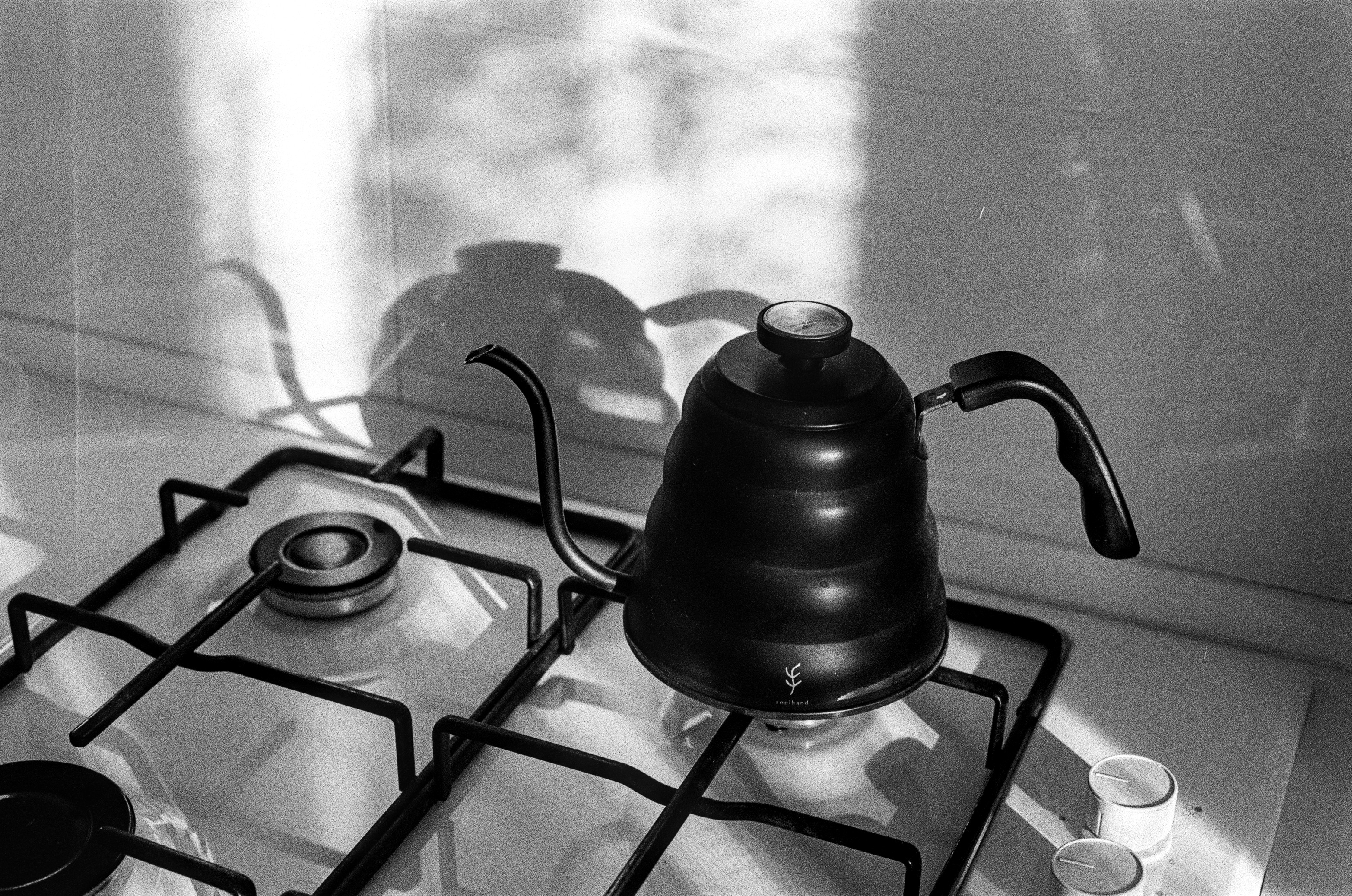 The image shows a black stovetop kettle. The kettle is made of metal and has a black finish. The kettle has a cone shape that tapers upward. Long, narrow spout in the shape of a goose's neck.
The kettle is lit by the sun and casts a shadow on the wall