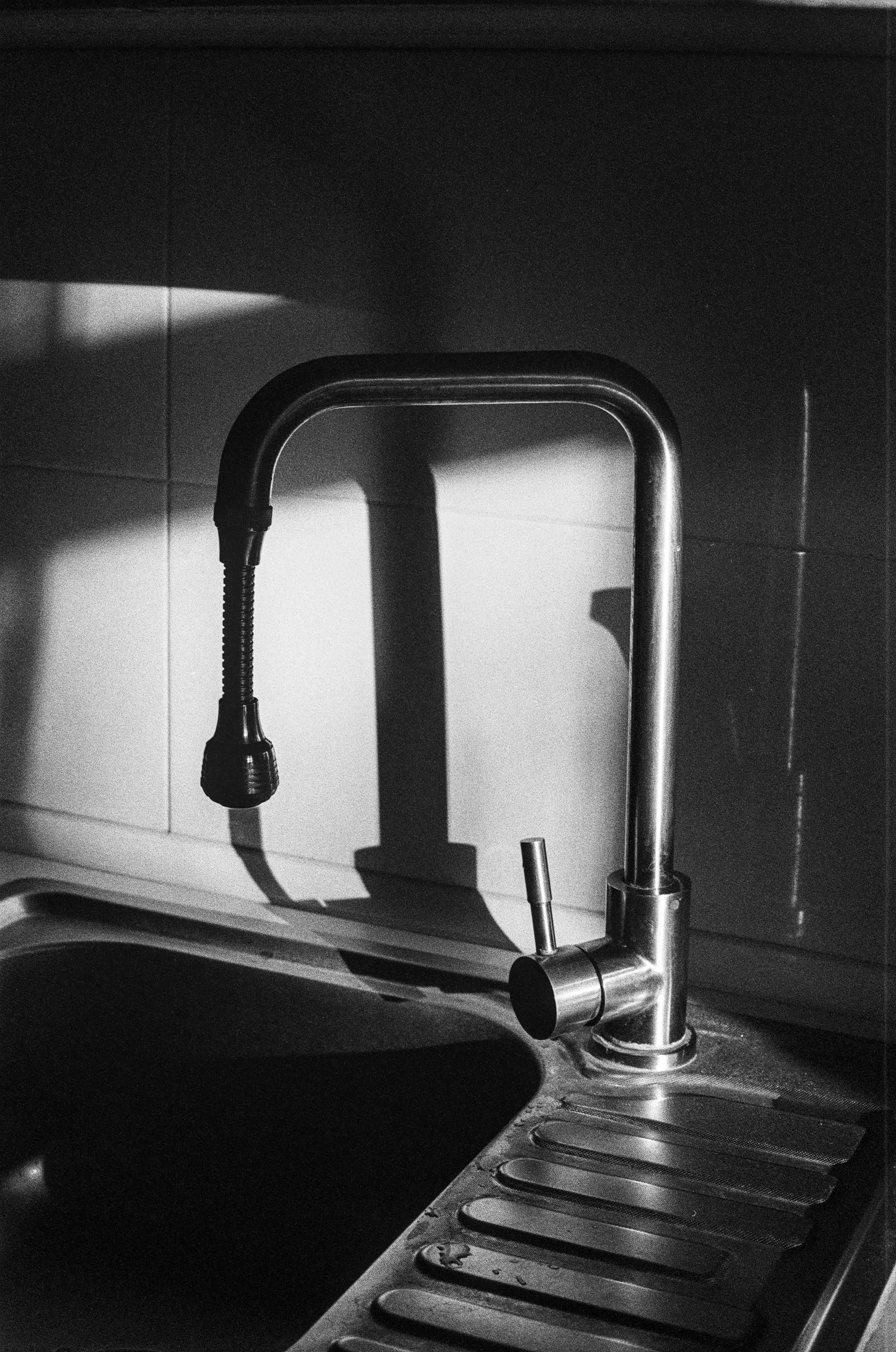 The image shows a black and white photo of a kitchen sink with a faucet on it. The sink is made of metal. It has a single basin and a drainboard. The faucet is also made of metal. The kitchen faucet is tall. It is lit by the sun and casts a shadow on the wall