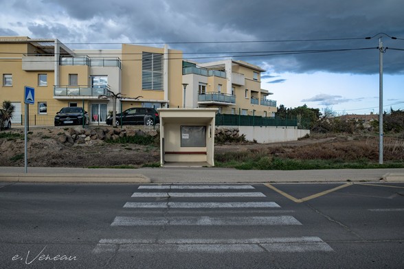 In front: a pedestrian crossing leading to a bus stop. Behind, a modern building with angular shapes, made of blocks with glass balconies, a parking lot with new city cars. On the left on the sidewalk is a blue sign indicating the pedestrian crossing. On the right an electric pole. The bus stop and the buildings are in yellow, white and gray shades. The cloudy sky is in almost blue hues.