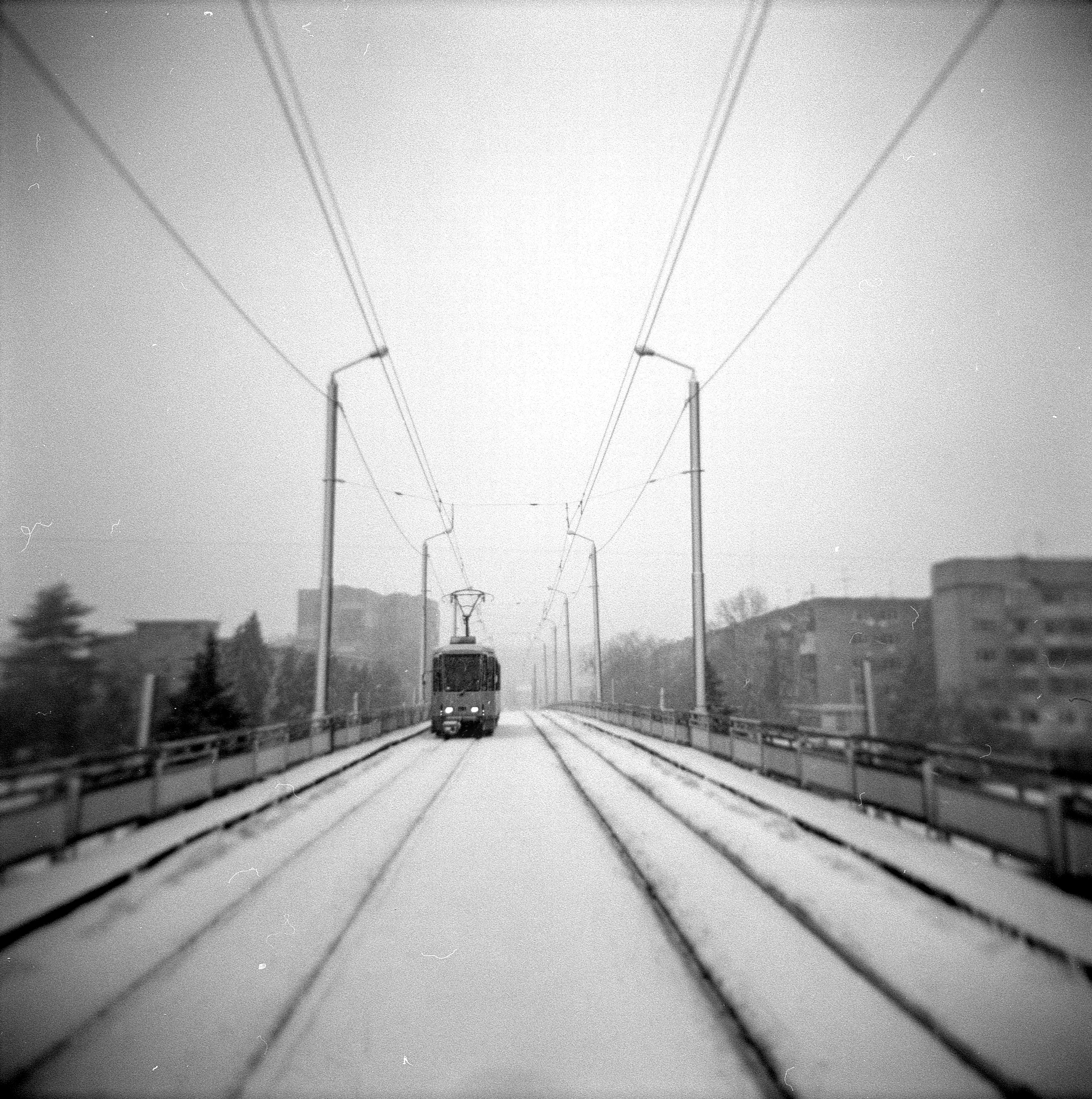 The image is a black and white photo of a tram on a bridge. The tram is in the middle of the bridge and moves toward the observer. The tracks and bridge are covered with snow.
In the background, houses are visible in the fog.
The lines of the tracks below and the electrical wires above form the perspective 
