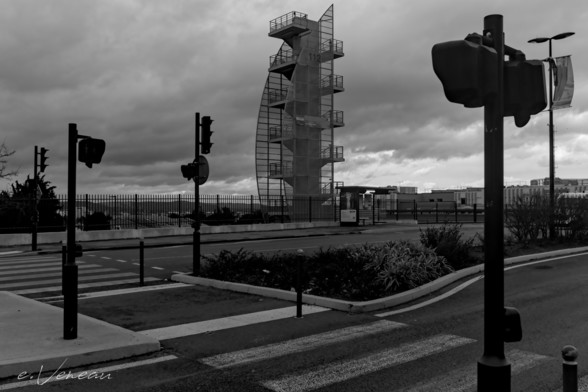 A double pedestrian crossing lined with traffic lights leads to a sidewalk with a barrier, a bus stop. Behind a tower (metal structure) of a fire center stands against a cloudy sky.