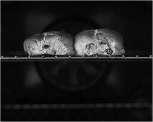 A black and white photograph of two conjoined hot cross buns, in an oven. Taken with a Nikkor 50mm f1.8D at eye level, in reflected window light at -2 stops.