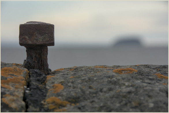 A colour photograph of a rusting iron pin and nut set in the black mortar joint of a stonewall. De-focused in the middle of the frame is the Bristol Channel at high tide and in the backdrop is Steepholm Island.