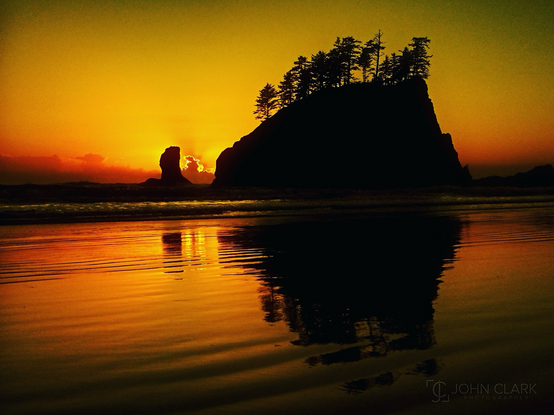 Dark rock with trees at the top with sunset oranges in the sky and reflecting on a low tide ocean water