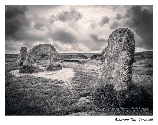 Black and white photo of an upright stone with a large hole through it flanked by two standing stones, with a dramatic cloudscape behind.