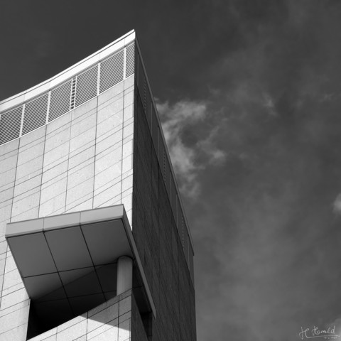 Square black and white image of the upper edge of a high building under a dark sky with some feathery clouds. No windows but one large balcony with a roof sticking out of the fassade is visible.