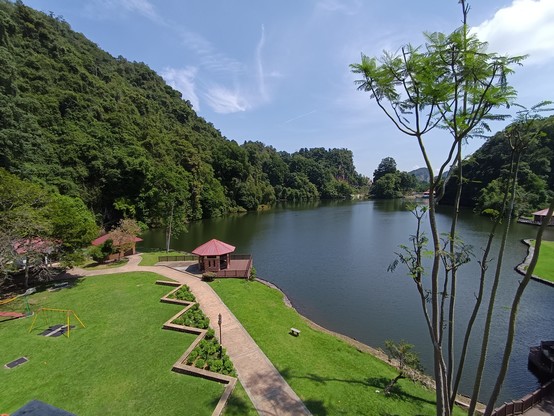 A shot of the lake from an elevated point. The lake is in the background. The foreground shows a walking path along the edge of the lake with a shelter near the middle.